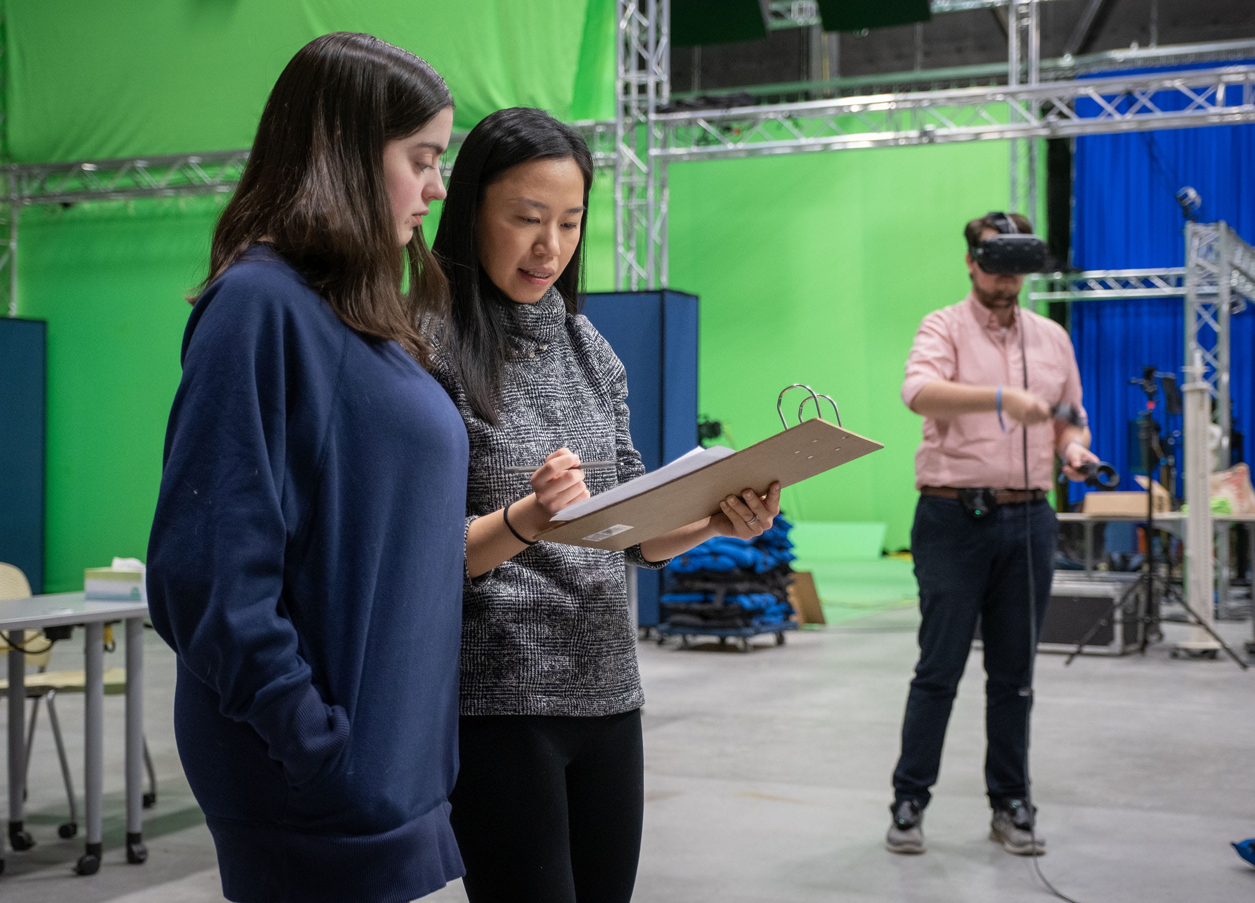Two female students looking at a clipboard while a male student uses a VR headset